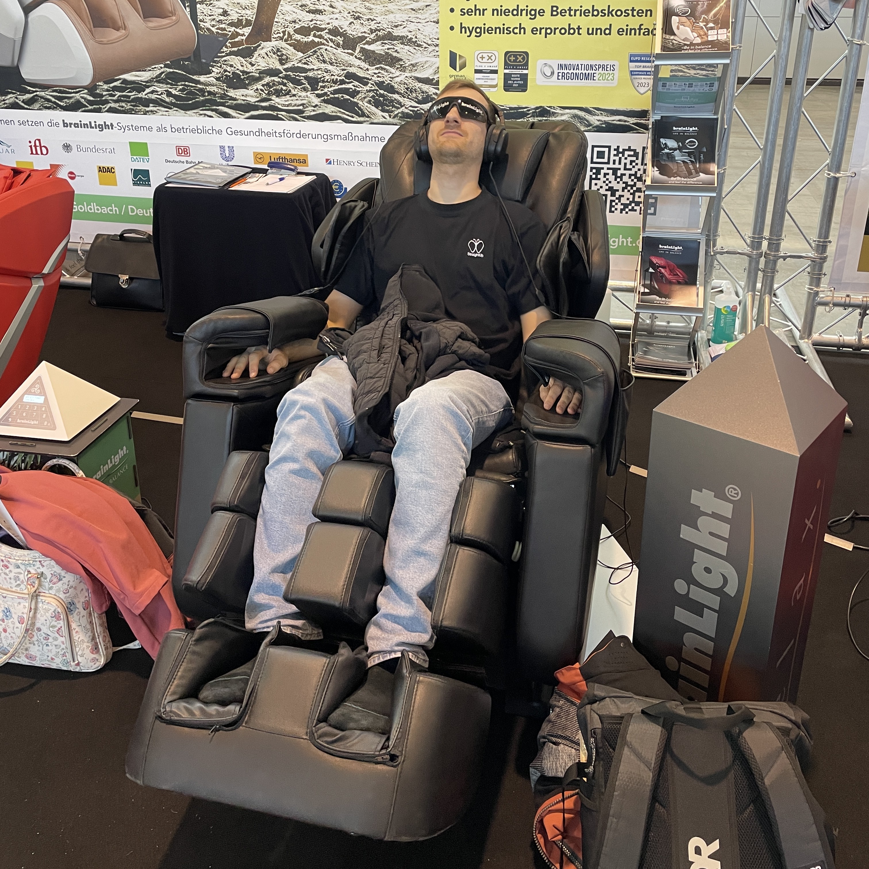 Eine Person sitzt entspannt in einem hochwertigen Massagesessel auf der Therapro-Messe, trägt Kopfhörer und eine Sonnenbrille. Im Hintergrund sind Messestände und ein großes Werbebanner zu sehen, das Informationen zu Gesundheits- und Wellnesssystemen zeigt.