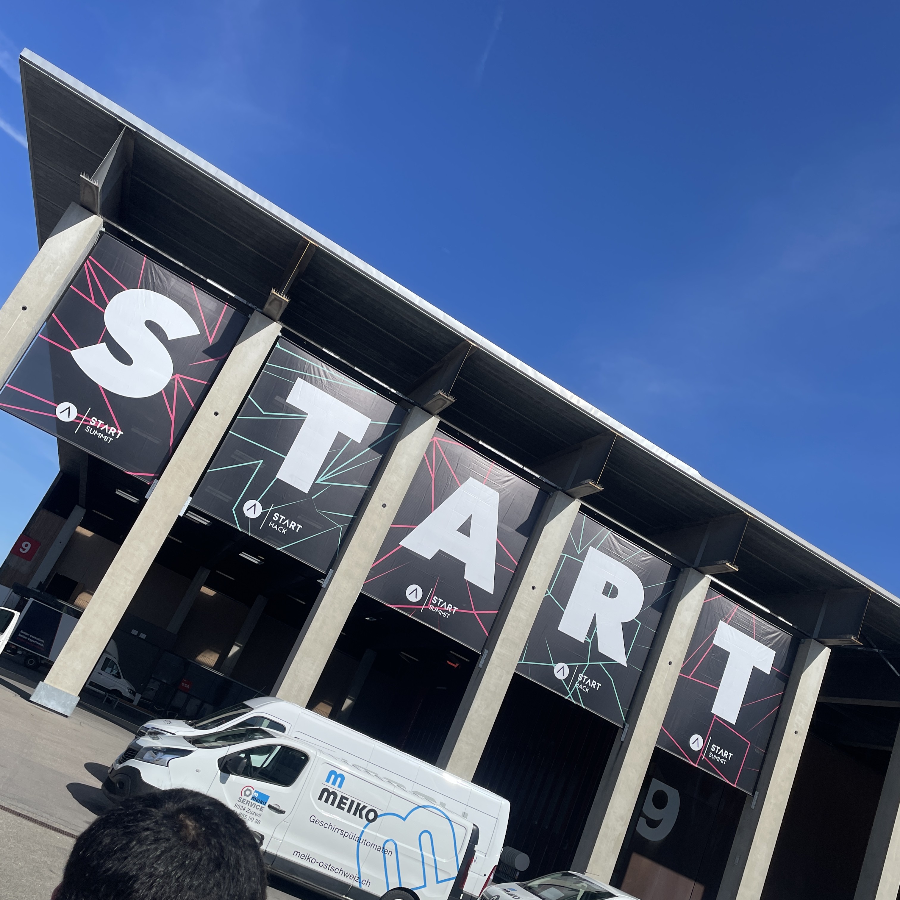 Außenaufnahme des START Summit 2025 in St. Gallen, mit riesigen Bannern an einem Halleneingang, die das Wort „START“ bilden. Der Himmel ist klar und blau.